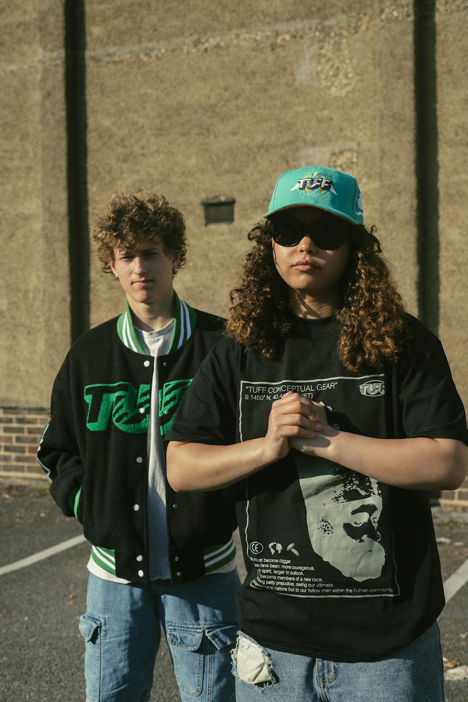 Zwei Personen in Streetwear stehen vor einer Betonwand.